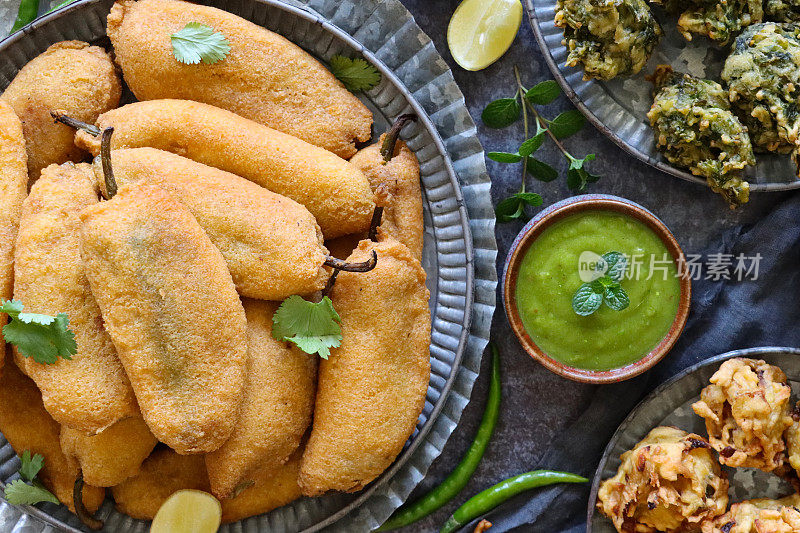 近距离的图片，托盘上有图案的自制绿辣椒油饼(mirch pakora)，盘子里的脆洋葱bhajis和palak pakora(菠菜油饼)，碗里的薄荷和香菜酸辣酱，印度菜素食小吃，灰色的背景，高架视图